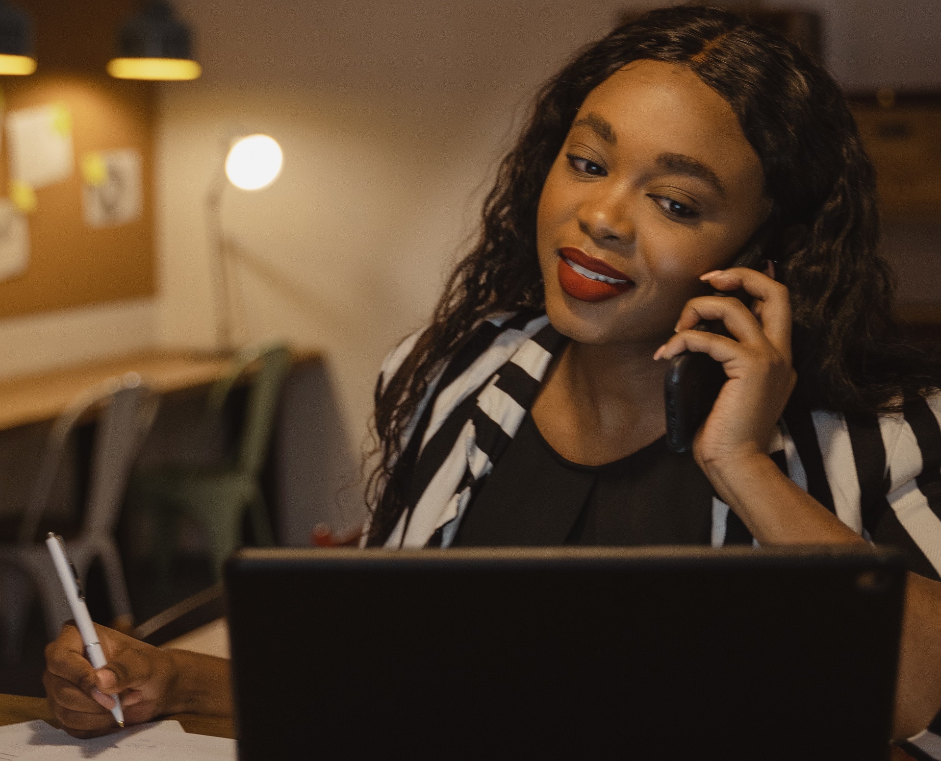 woman writing while on the phone looking at a computer